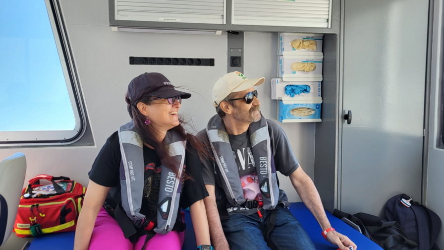 Ein Paar sitzt in einer Kabine in einem Boot. Beide tragen Rettungswesten. Sie schauen in die Ferne und lächeln.