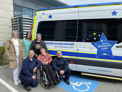 Sechs Personen stehen und sitzen vor einem Fahrzeug mit der Aufschrift „Wunschfahrt“. Das Fahrzeug hat eine bunte Gestaltung mit Sternen. Im Hintergrund ist ein modernes Gebäude sichtbar. Alle Personen wirken freundlich und lächeln.