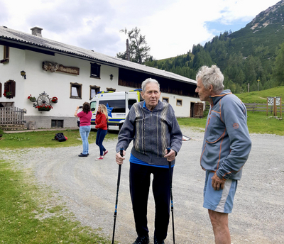 Zwei ältere Männer stehen im Freien und unterhalten sich vor einer Berghütte.