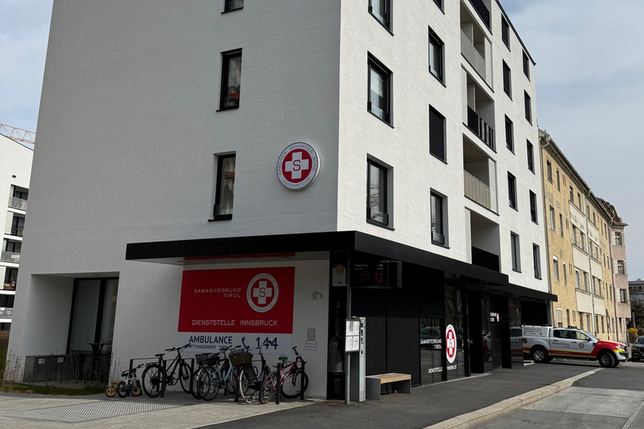 Ein modernes, mehrstöckiges Gebäude mit weißer Fassade. Im Erdgeschoss befindet sich ein Eingang mit einem roten Kreuz-Symbol. Vor dem Gebäude stehen mehrere Fahrräder. Der Himmel ist bewölkt.