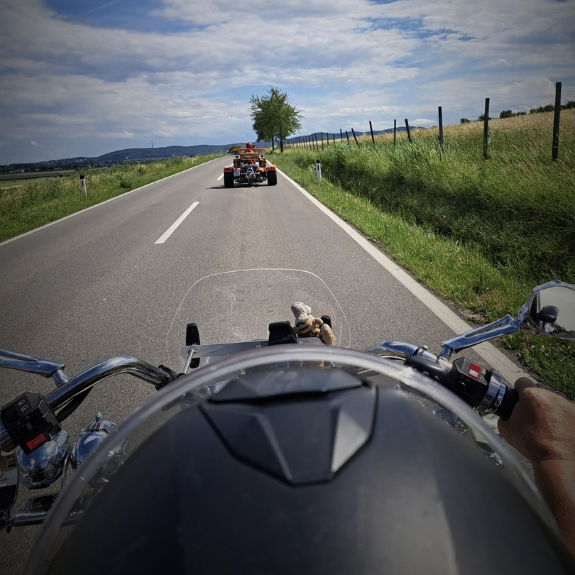 Blick aus der Perspektive eines Motorradfahrers auf eine Landstraße. Im Vordergrund sind das Motorrad und die Hände des Fahrers sichtbar. Auf der Straße fährt ein weiteres Fahrzeug, während im Hintergrund Bäume und ein klarer Himmel zu sehen sind.