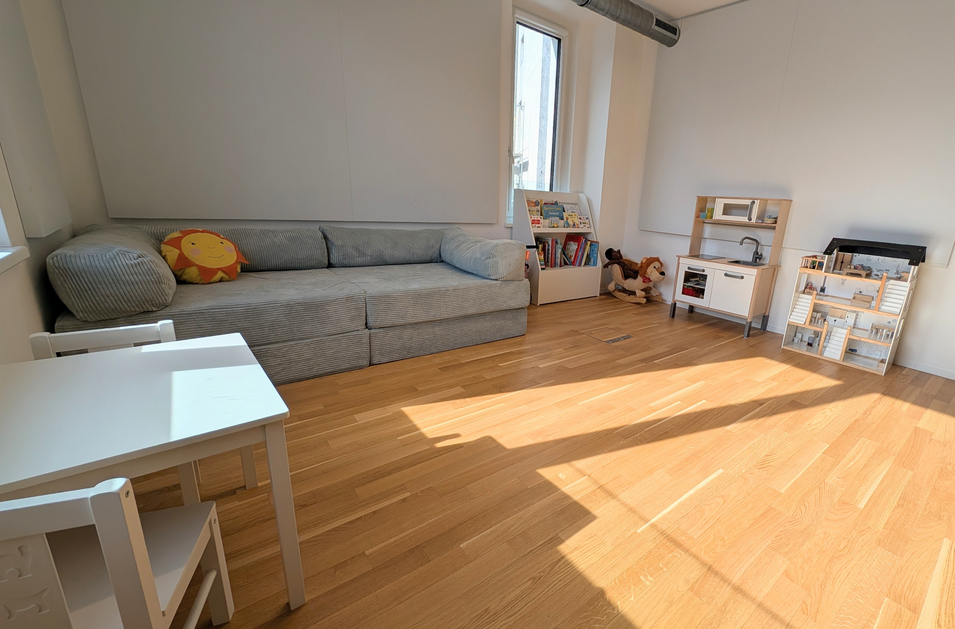 Ein heller Raum mit Holzfußboden. Links steht ein graues Sofa mit einem Kissen. Rechts ist eine kleine Spielküche, ein Bücherregal und ein Spielzeughund. In der Mitte steht ein kleiner Tisch mit zwei Stühlen. Ein Fenster sorgt für natürliche Beleuchtung.