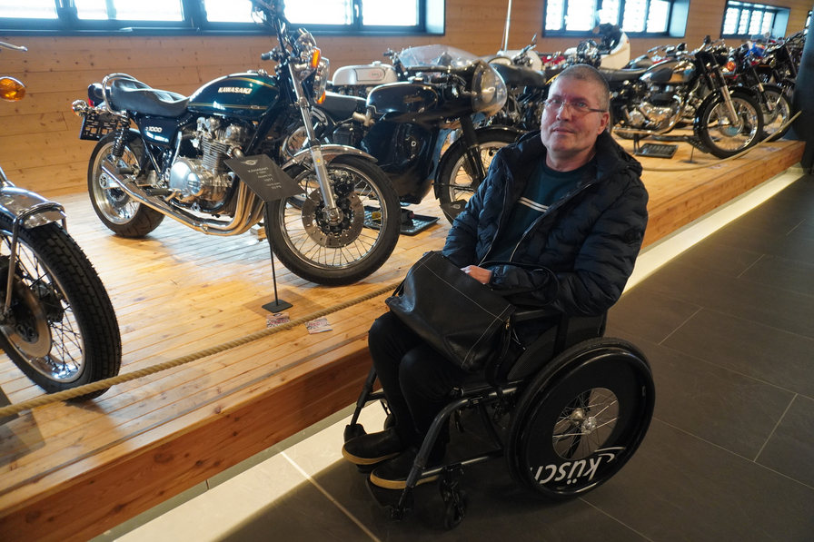 Ein Mann im Rollstuhl sitzt neben mehreren Motorrädern in einem Museum.