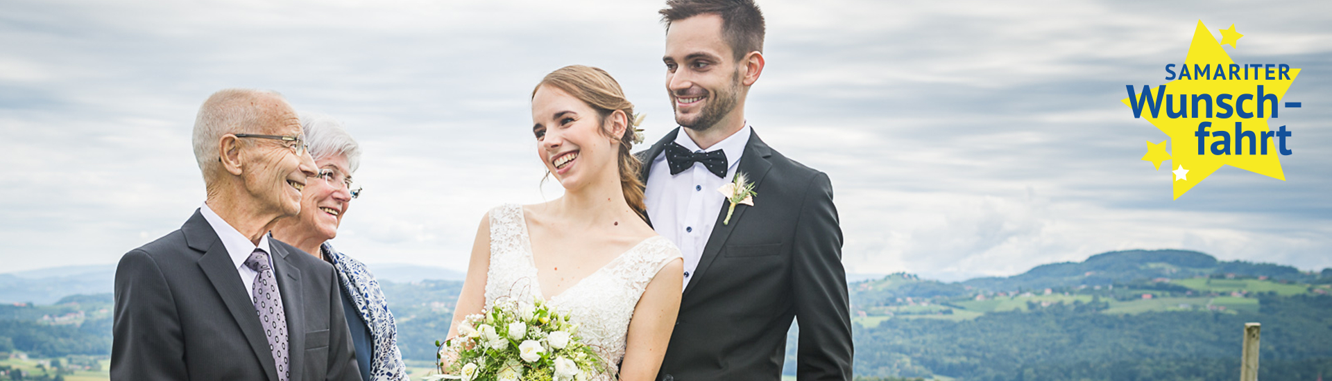Eine Braut in einem weißen Kleid mit Blumenstrauß steht lächelnd neben einem Bräutigam in einem schwarzen Anzug. Neben ihnen stehen zwei ältere Personen, die ebenfalls lächeln. Im Hintergrund sind sanfte Hügel und ein bewölkter Himmel zu sehen.