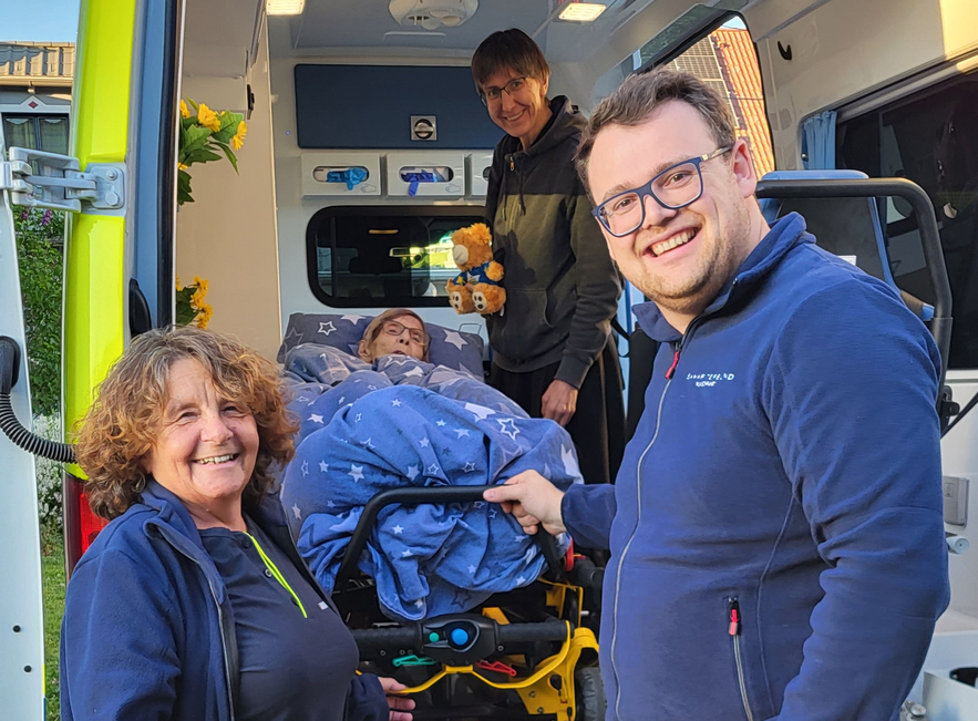 Eine Krankenschwester und ein Rettungssanitäter stehen an einem Krankenwagen. Der Sanitäter lächelt, während sie eine Trage mit einer Person darauf ins Fahrzeug schieben. Im Hintergrund ist ein weiterer Rettungsdienstmitarbeiter sichtbar. Die Szene vermittelt ein Gefühl von Unterstützung und Fürsorge.