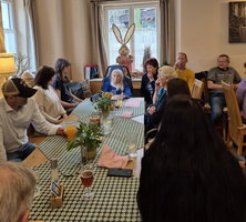 Eine Gruppe von Menschen sitzt an einem Tisch in einem gemütlichen Raum. Im Hintergrund steht eine große, dekorative Osterfigur eines Hasen. Es sind Getränke und Pflanzen auf dem Tisch, und die Teilnehmer scheinen an einer Unterhaltung oder Feier teilzunehmen.